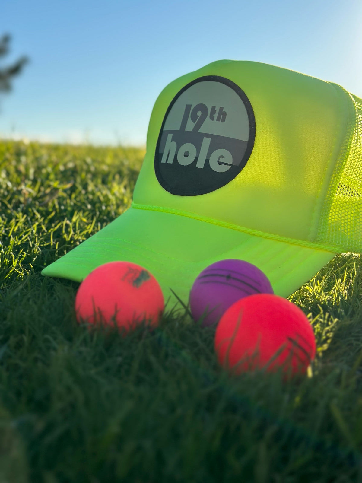 Neon yellow 19th hole golf trucker hat with golf balls in grass.