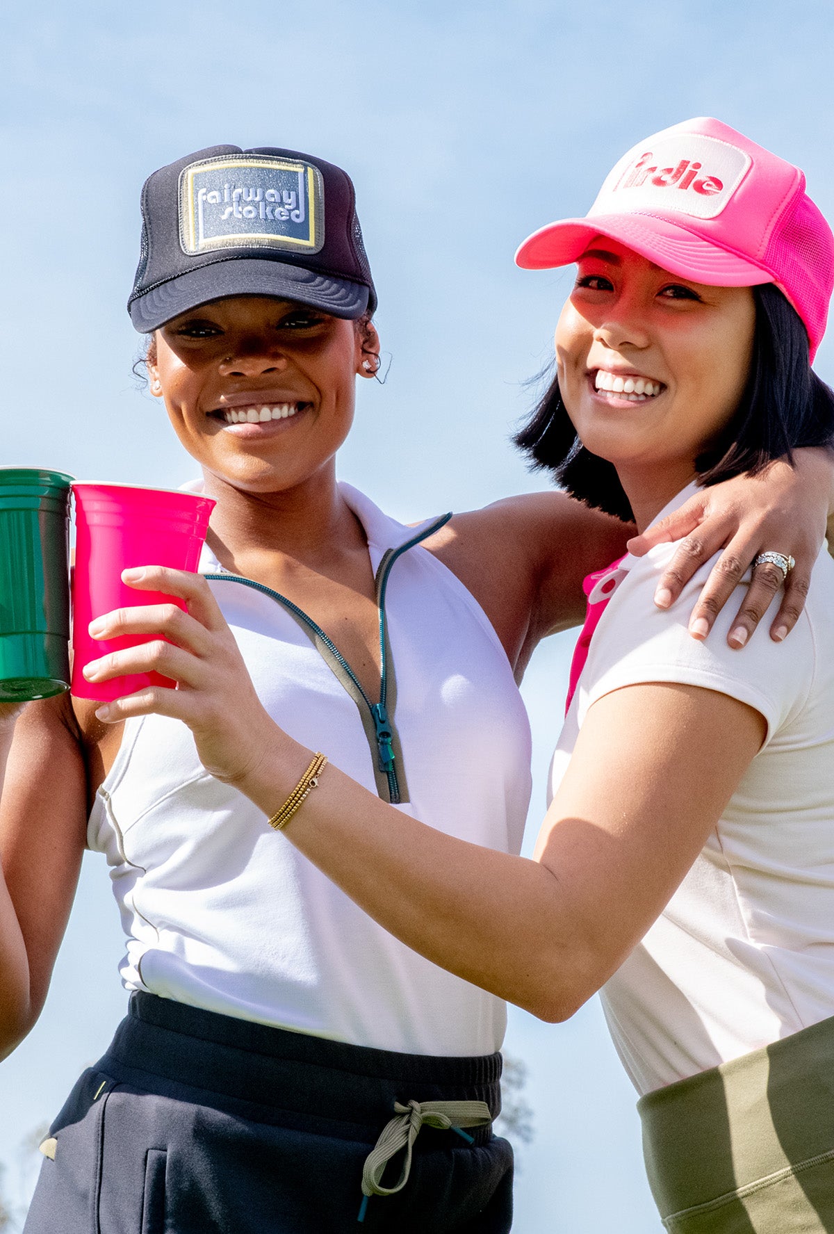 Two model cheers-ing with cups.  One is wearing a black Fairway Stoked golf  trucker hat and the other is wearing a Hot Pink Pirdie golf trucker hat.